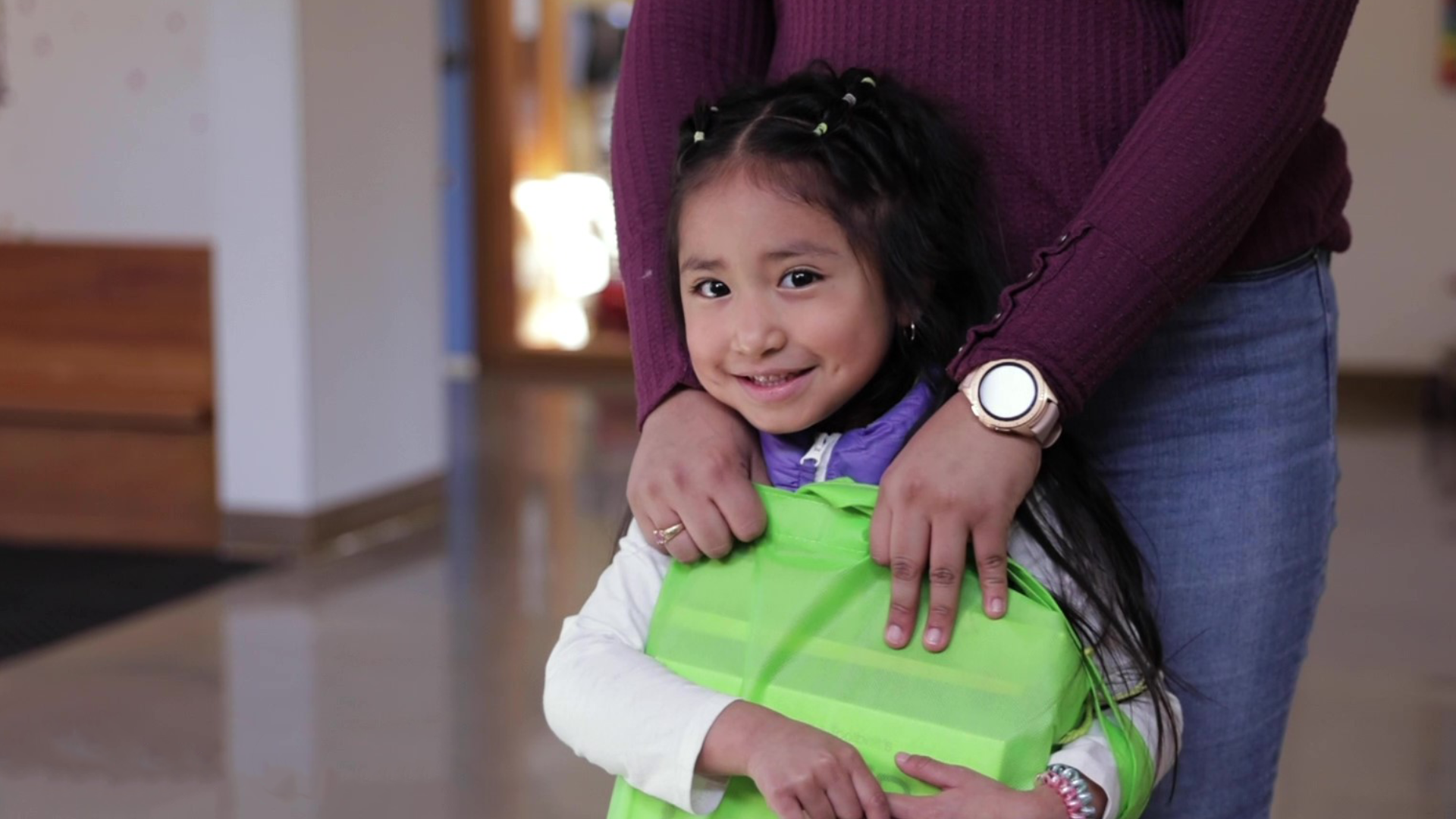 A child holds a green tote bag full of 14 books from the Book Bank. Her mother stands behind her with her arms around her. 